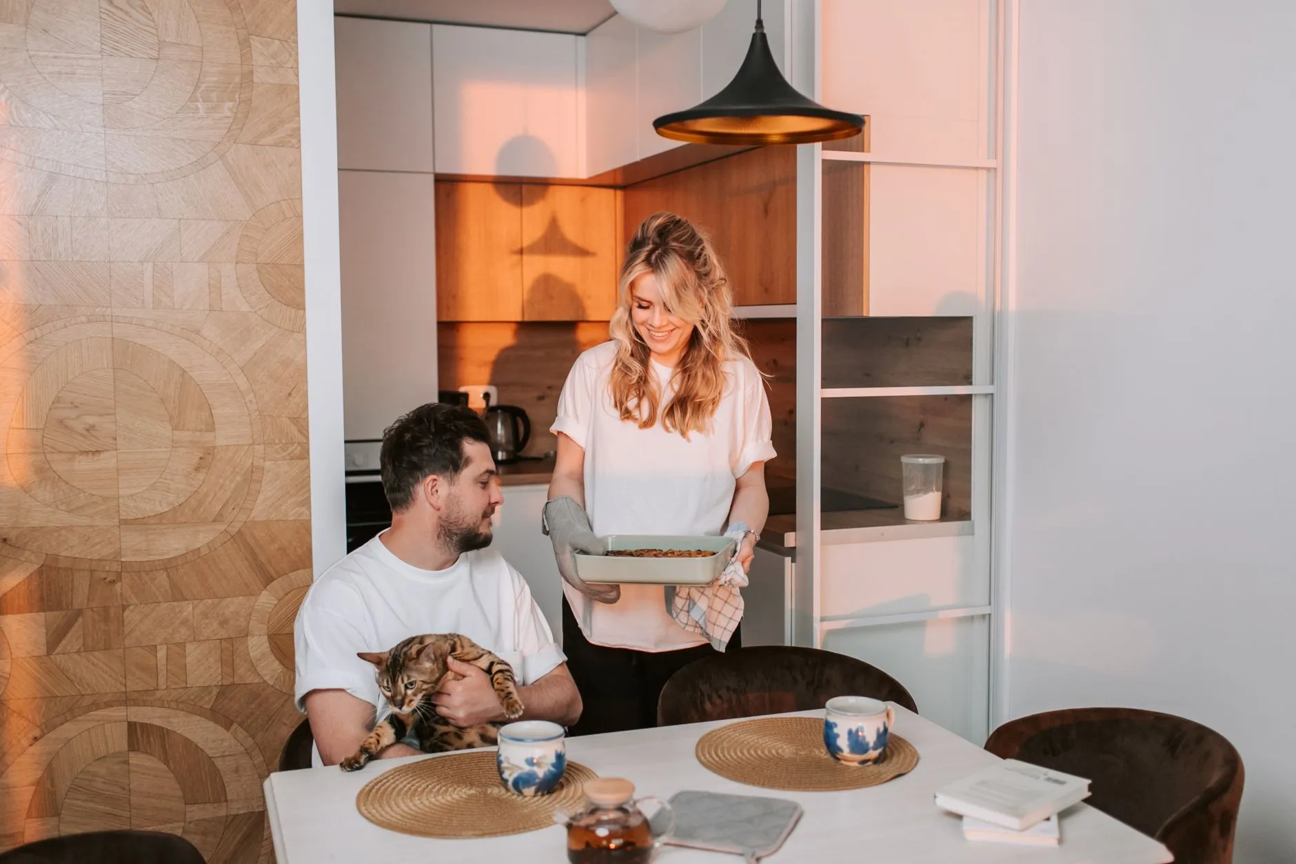 A joyful couple at home enjoying dinner with their pet cat in a cozy kitchen.