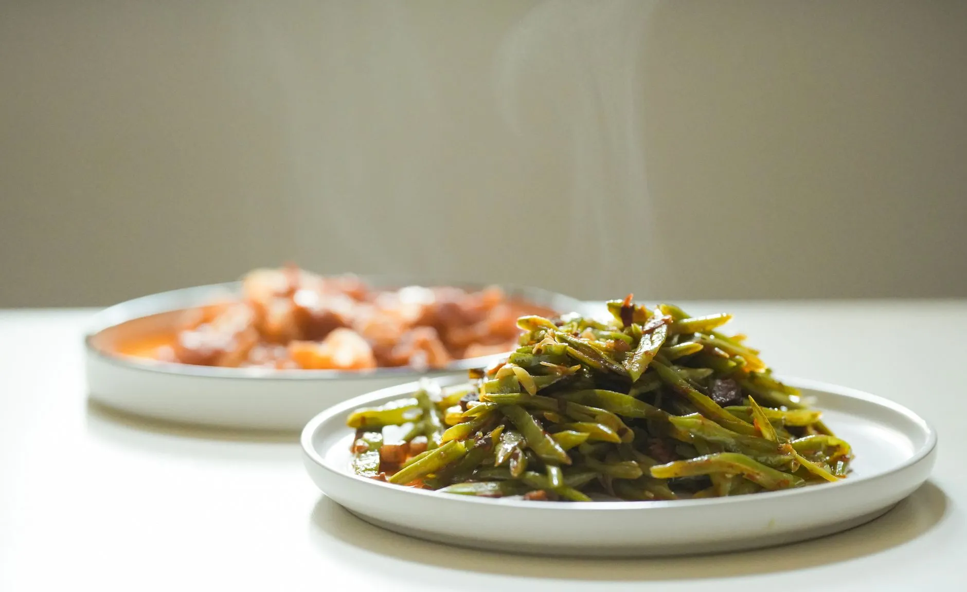 Close-up of a steaming green bean dish with another steaming stew in the background, served on white plates.