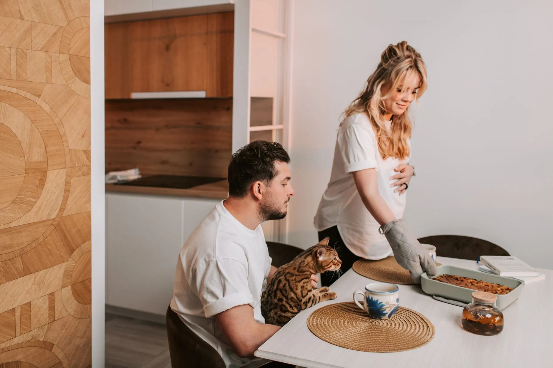 Expectant mother serves dinner while partner and cat sit nearby in a cozy dining space.