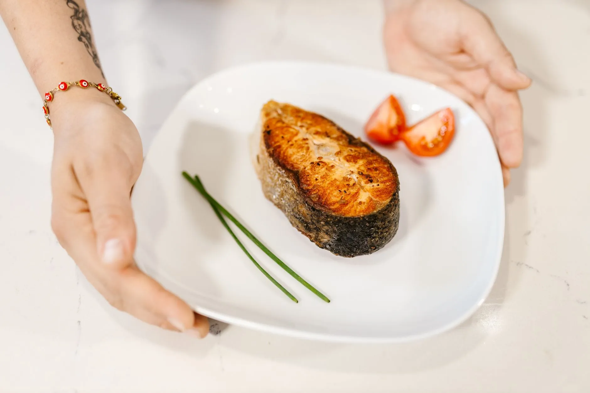 Close-up of grilled salmon served with tomatoes and chives on a plate, held by tattooed hands.
