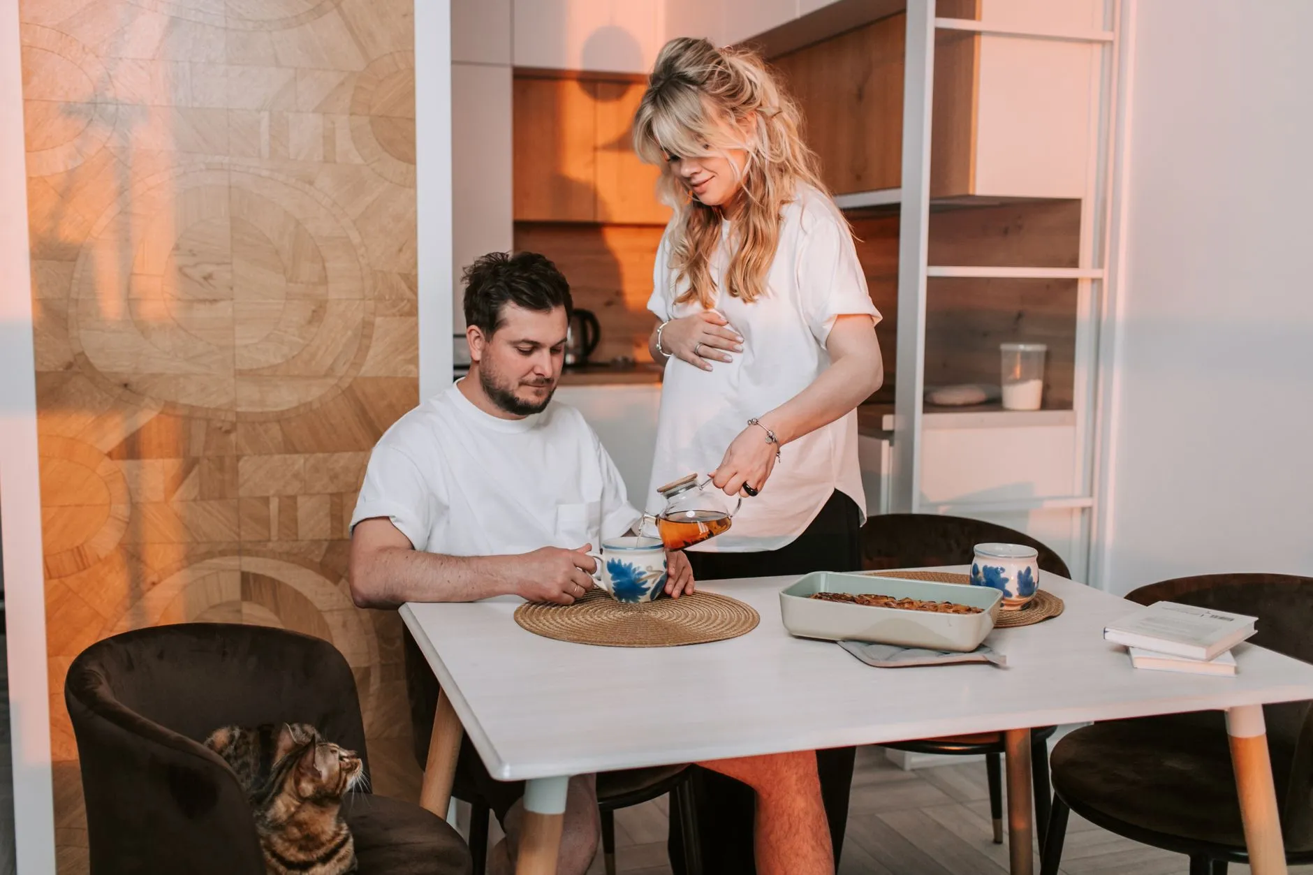 A pregnant woman and her partner sharing a peaceful afternoon with tea and a cat.
