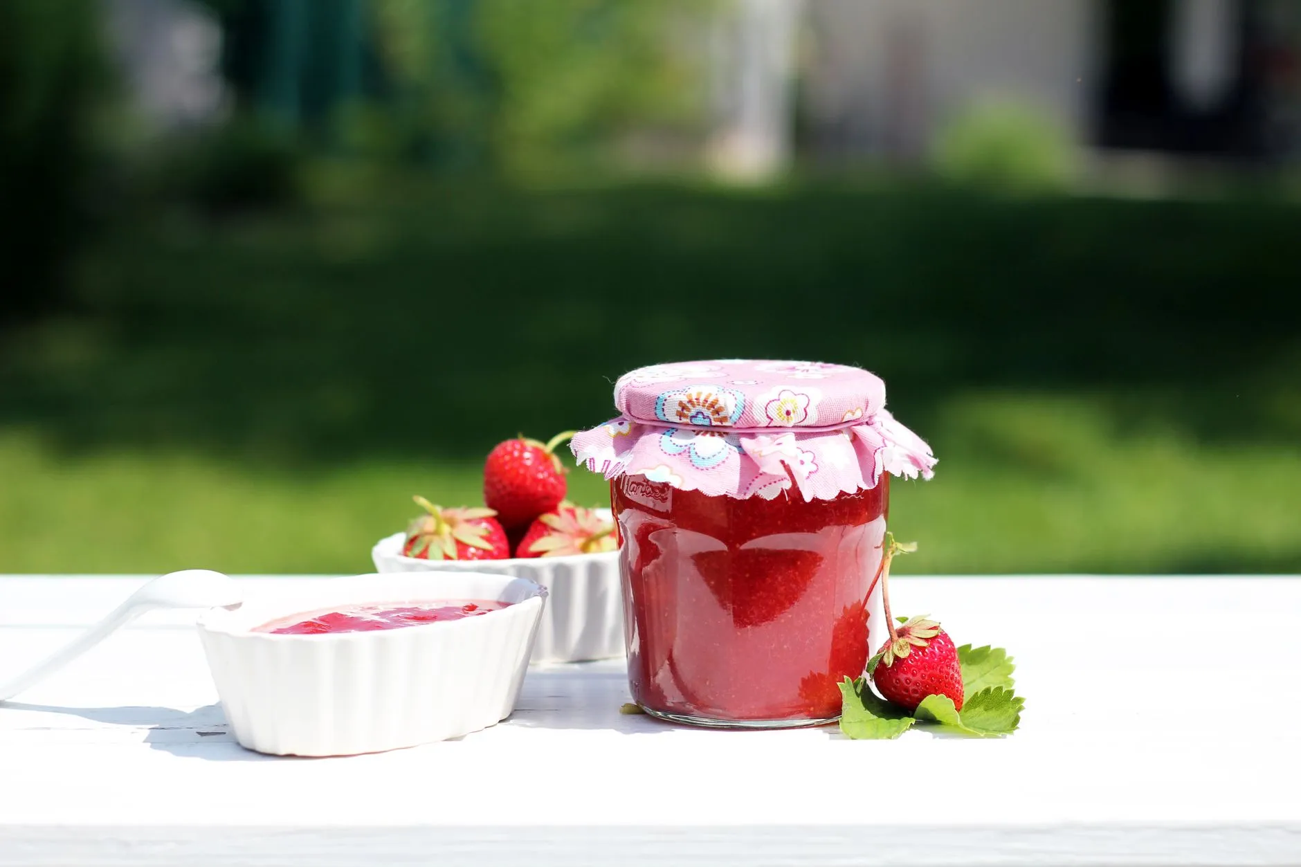 Fresh homemade strawberry jam in a decorative jar with ceramic bowls on a white table outdoors.