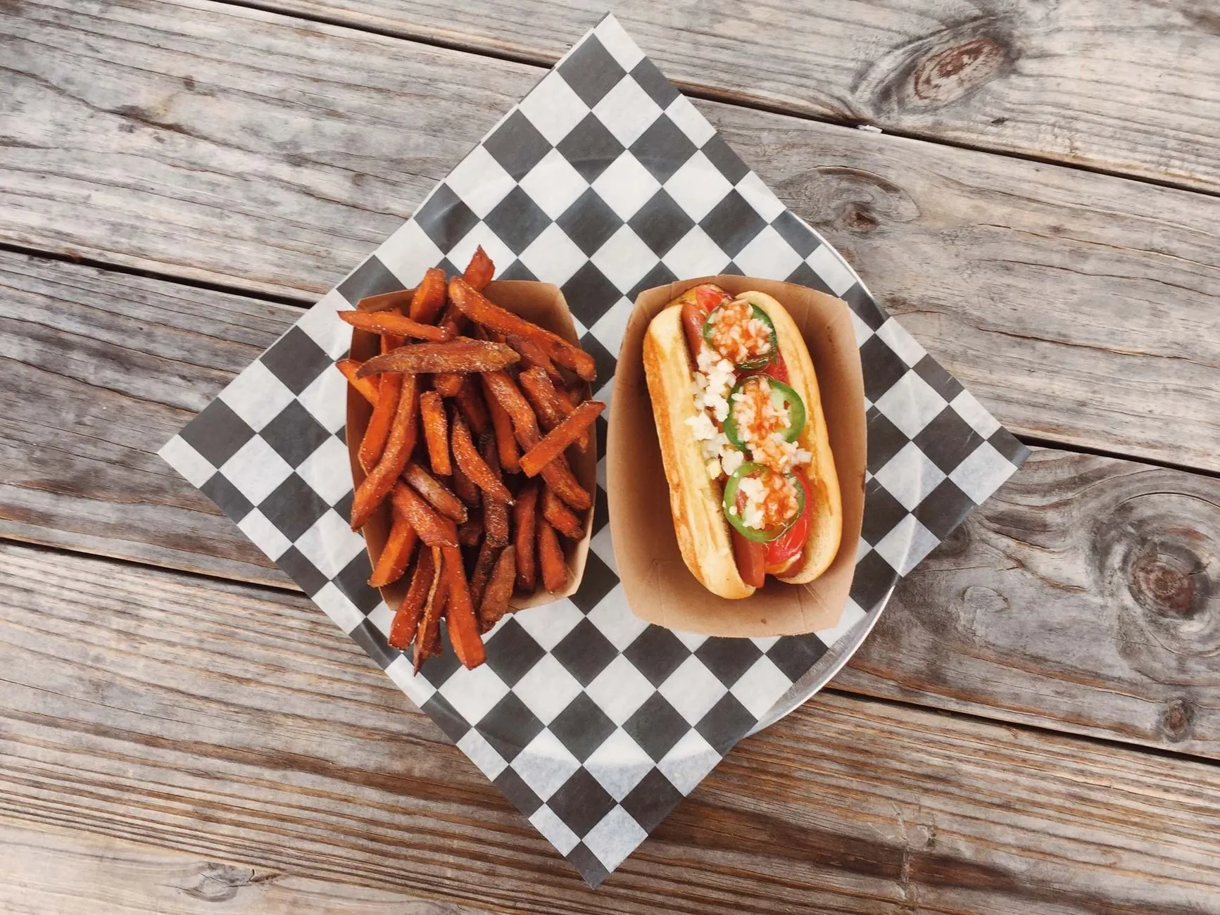 Flat lay of a gourmet hot dog with fresh toppings and sweet potato fries served on a rustic wooden table.